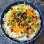A close-up of fluffy mashed potatoes topped with crispy bacon bits, shredded cheddar cheese, and chopped green onions in a blue bowl.