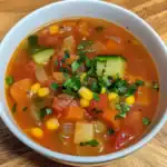 Savory vegetable soup featuring carrots, zucchini, corn, and tomatoes garnished with chopped herbs in a white bowl.