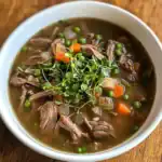 Hearty beef and vegetable soup with peas, carrots, and fresh herbs in a white bowl.