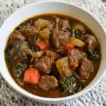 Hearty beef stew with tender beef chunks, carrots, potatoes, and kale in a flavorful broth, served in a white bowl.