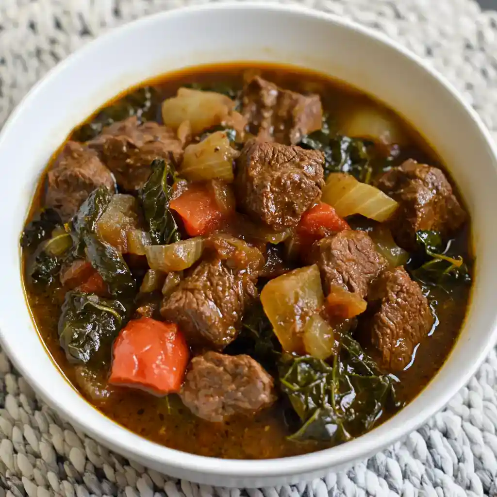 Hearty beef stew with tender beef chunks, carrots, potatoes, and kale in a flavorful broth, served in a white bowl.