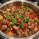Hearty beef stew with tender meat, garnished with fresh herbs and pomegranate seeds in a stainless steel pot.