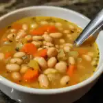 Hearty white bean and vegetable soup in a white bowl with a spoon, featuring carrots, celery, and herbs, perfect for a comforting meal.